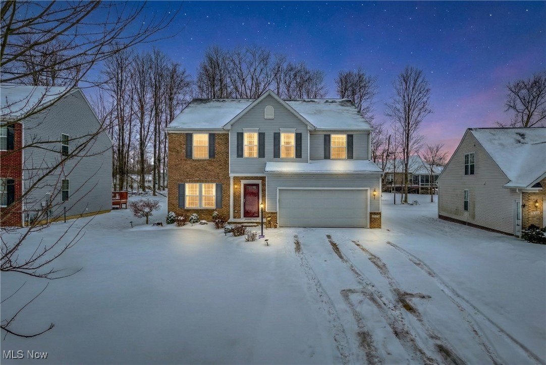 Two-story brick and gray-sided home with a two-car garage, warmly lit windows, and snow-covered grounds.