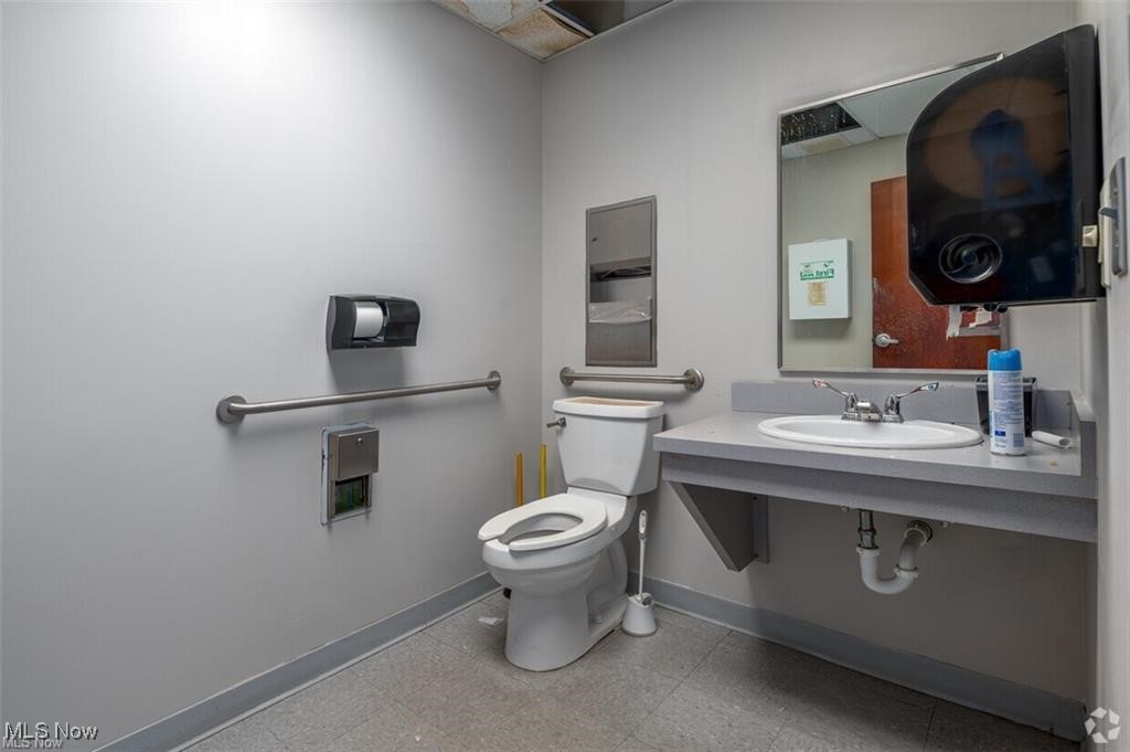 Bathroom with a toilet, grab bars, and a wall-mounted sink beneath a mirror.