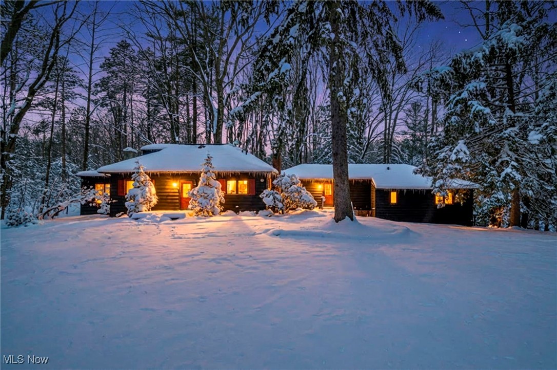 Snow-covered log home glowing with warm window light in a wooded setting at dusk.