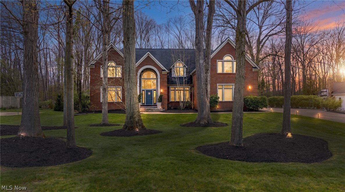 Two-story brick home with an arched entrance and glowing windows at dusk, framed by mature trees.