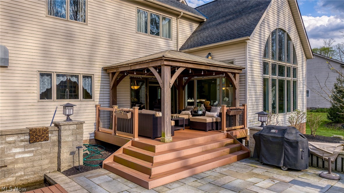 Rear of a beige home with a covered, furnished wooden deck and stone-paver patio.