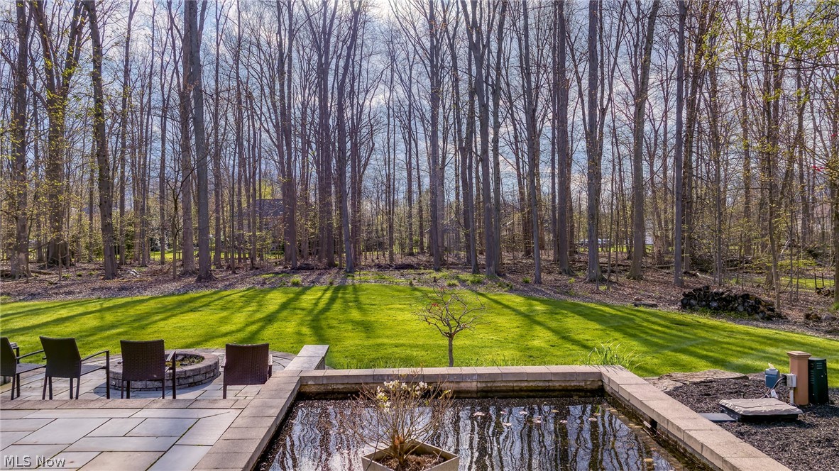 Wooded backyard with a green lawn, stone patio seating, and a reflecting pond.