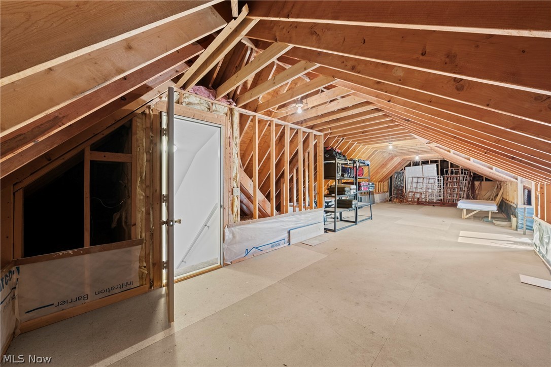 Unfinished attic with exposed roof framing, a plywood floor, and shelving at the far end.