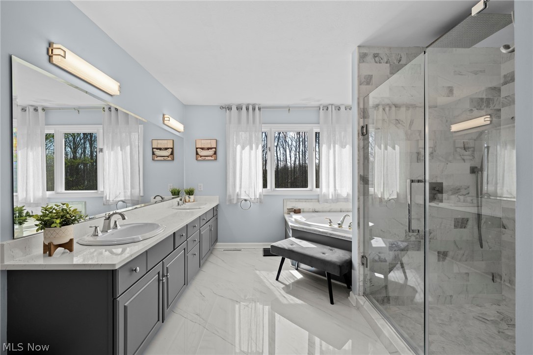Primary bathroom with gray double vanity, marble shower, soaking tub, and windows overlooking leafless trees.