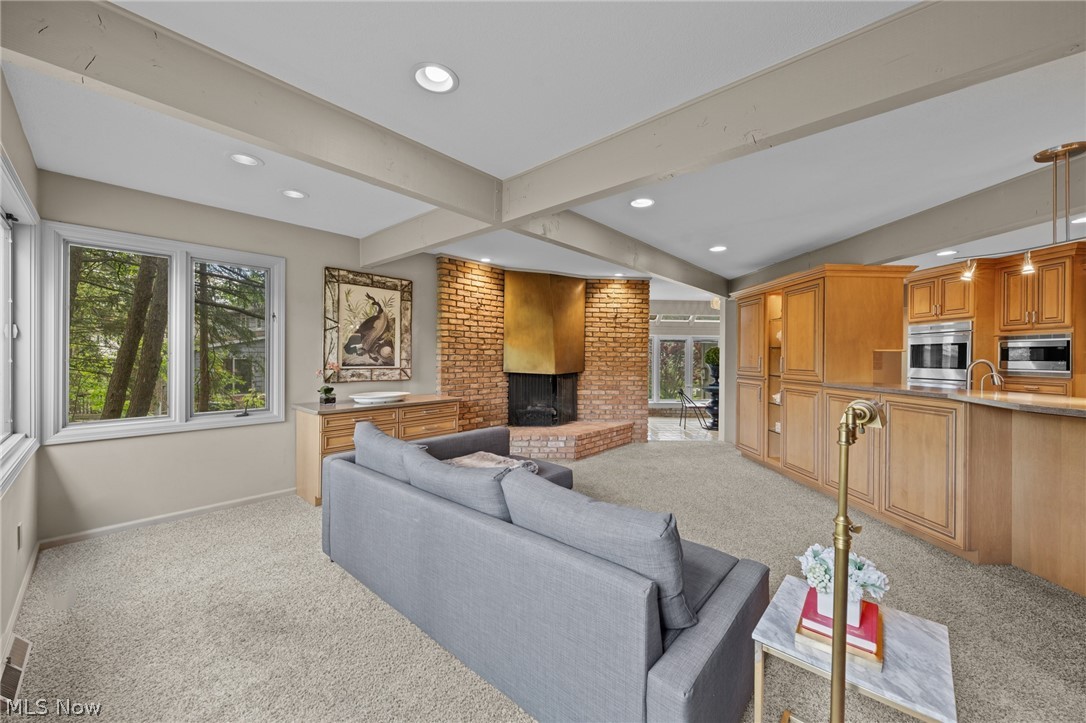 Living room with a gray sofa, brick fireplace, vaulted beamed ceiling, and adjoining wood-cabinet kitchen.