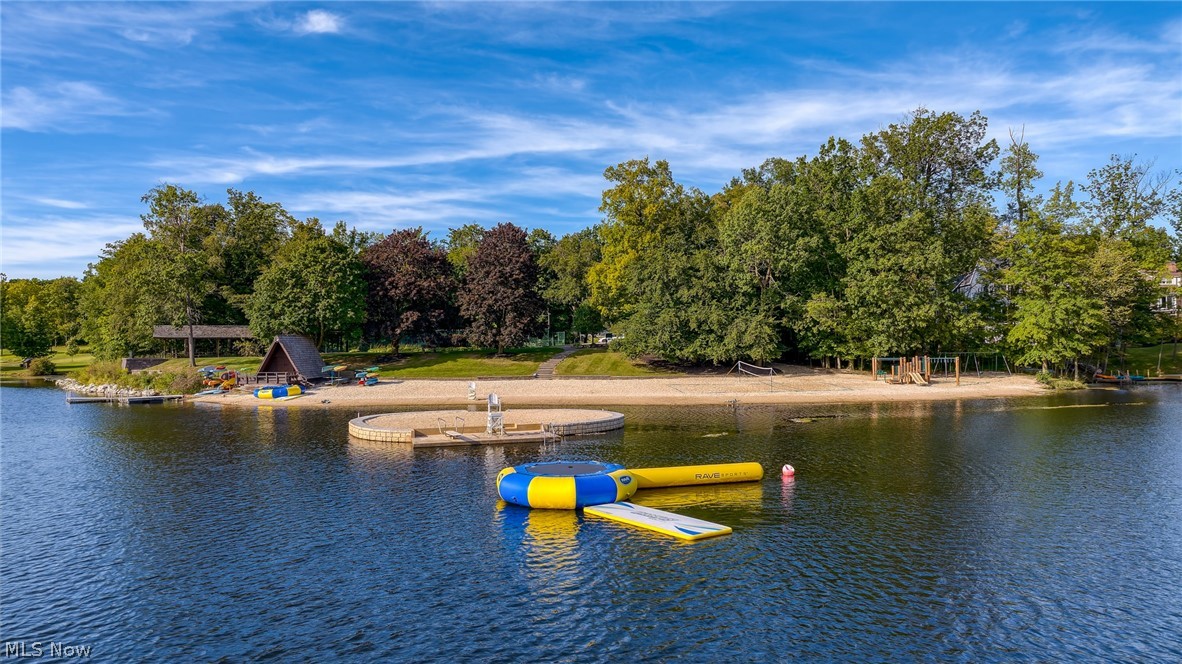 Lakefront beach with floating inflatables, a circular swimming platform, and trees along the shore.