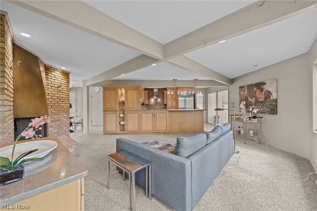 Living room with gray sofa, brick fireplace, and open kitchen beneath exposed ceiling beams.