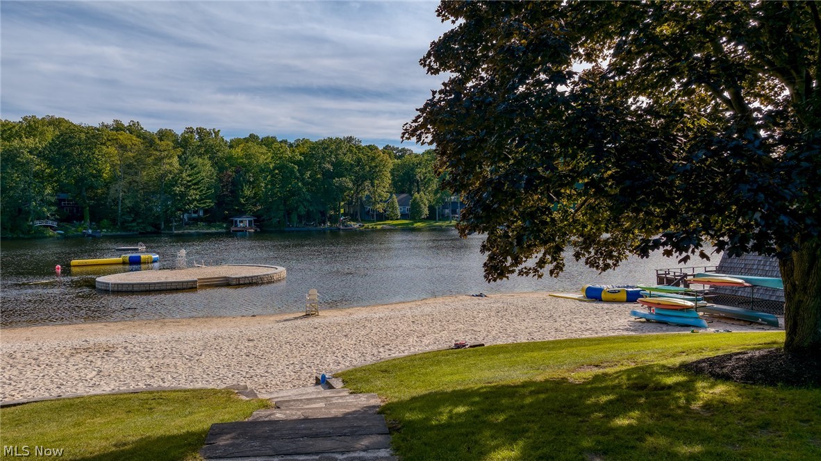 Sandy lakeside beach with a floating dock, colorful kayaks, and wooded homes across the water.