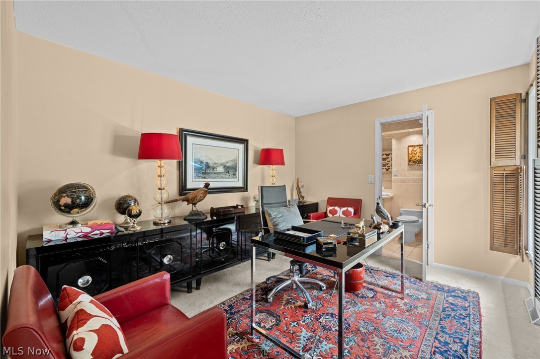 Furnished home office with a glass desk, red leather chairs, and patterned rug beside an open bathroom doorway.