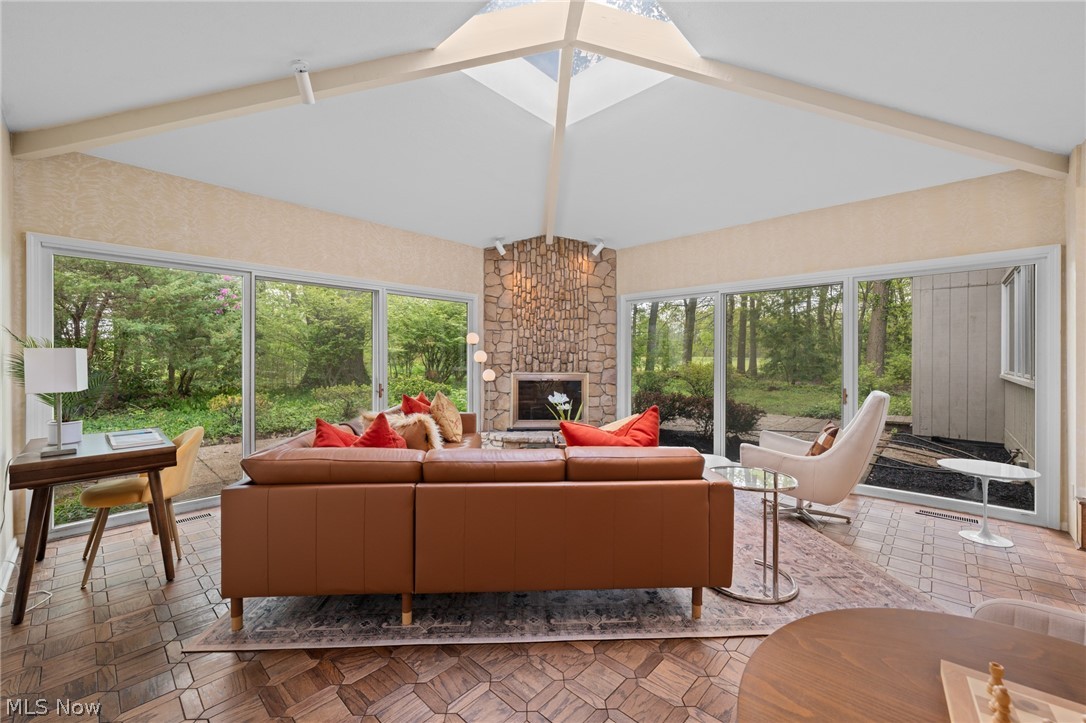 Sunroom with a stone fireplace, vaulted ceiling, and broad windows overlooking wooded grounds.