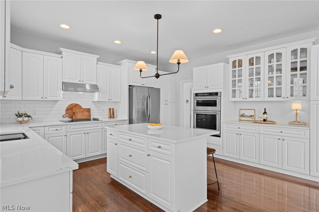 White kitchen with a large island, glass-front cabinets, stainless steel appliances, and warm wood floors.