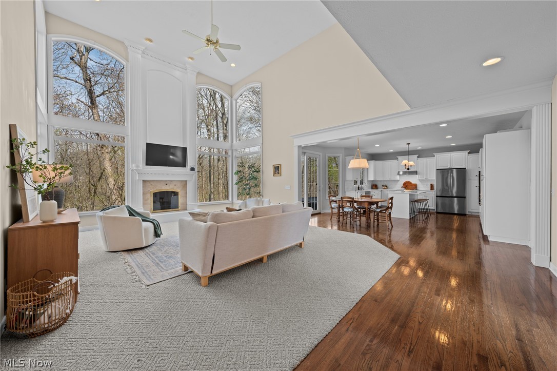 Living room with vaulted ceilings, a stone fireplace, and tall windows overlooking trees, open to the kitchen.