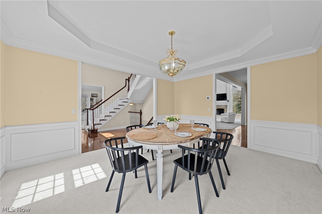 Dining room with a round table and black chairs beneath a gold chandelier, beside a staircase.