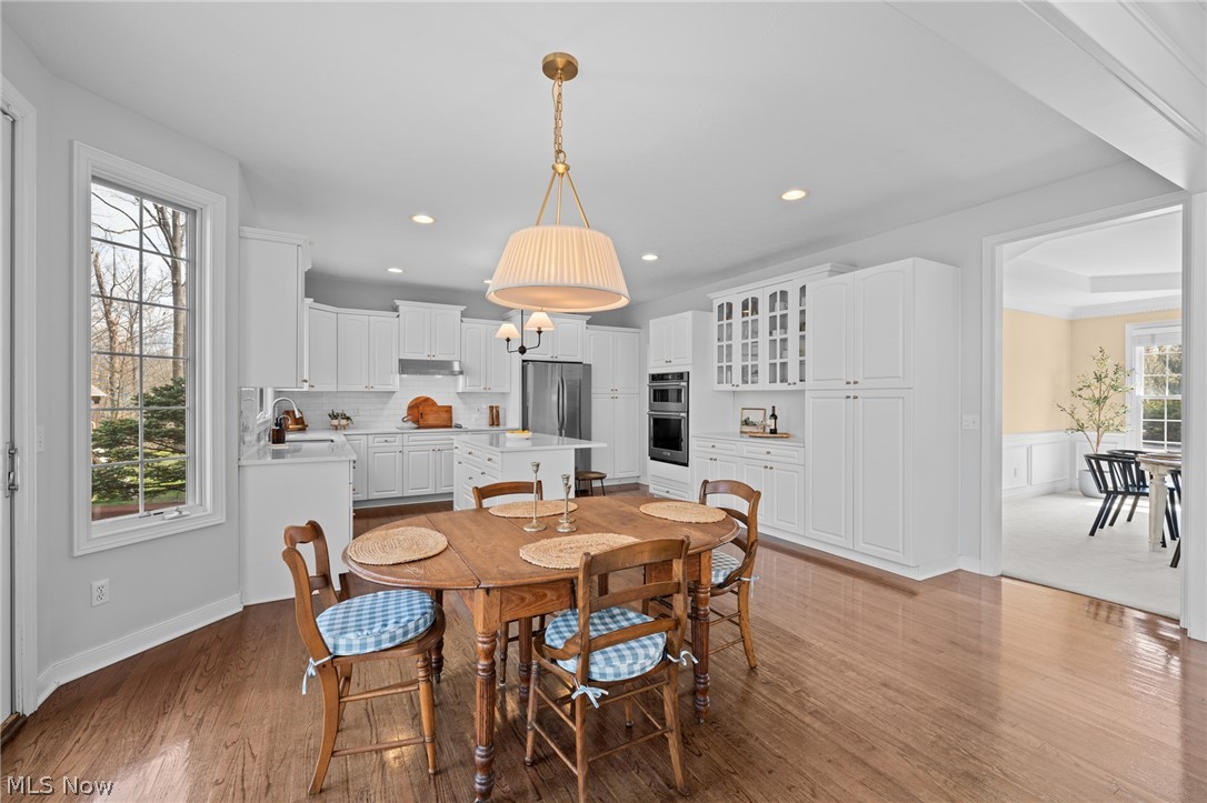 White kitchen with stainless steel appliances, central island, and wooden dining table beside a large window.