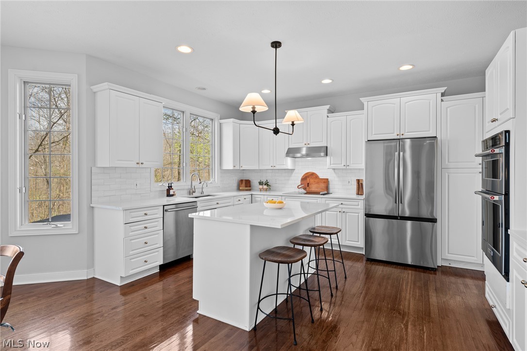 White kitchen with a large island, three stools, stainless steel appliances, and dark hardwood floors.
