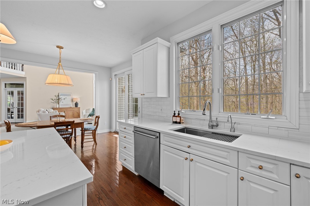 White kitchen with a double sink beneath large windows overlooking leafless trees, opening to a dining area.