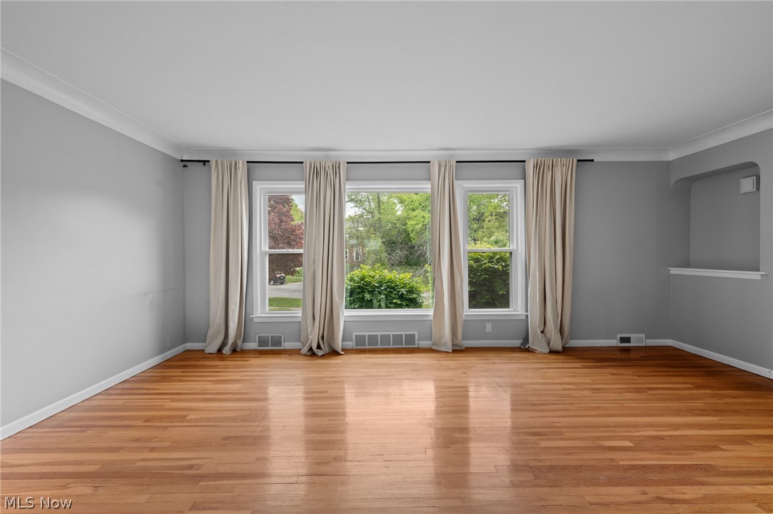 Vacant living room with hardwood floors, gray walls, and three large curtained windows.