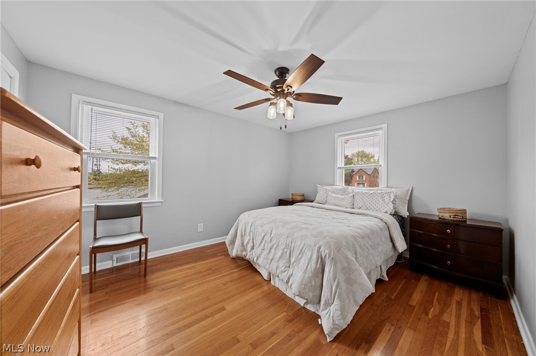 Bedroom with hardwood floors, a white bed, two windows, and a wood ceiling fan.