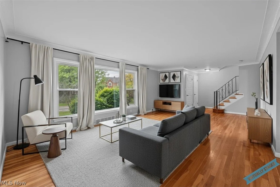 Living room with a gray sofa, broad curtained windows, hardwood floors, and a staircase.