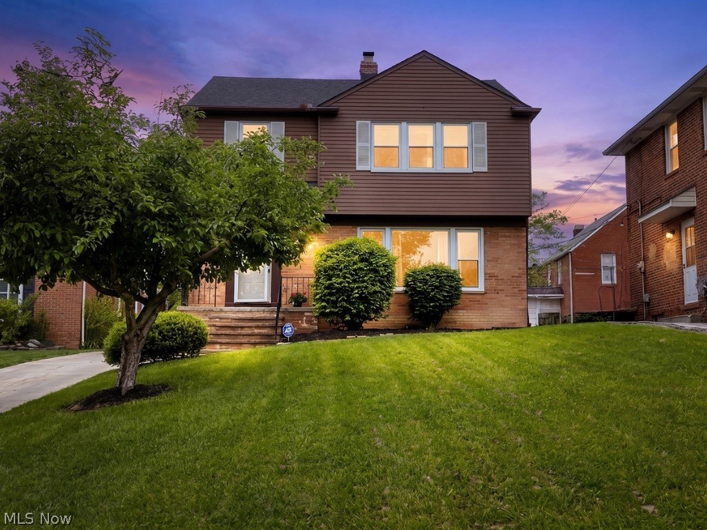 Two-story brick-and-siding home with warmly lit windows, a leafy tree, and front lawn at dusk.