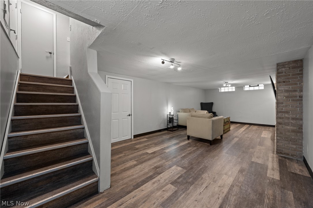 Finished basement living area with a beige sofa, wood-look flooring, and stairs beside a brick column.