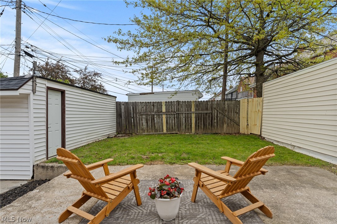 Fenced backyard patio with two wooden chairs and a potted flowering plant beneath a large tree.
