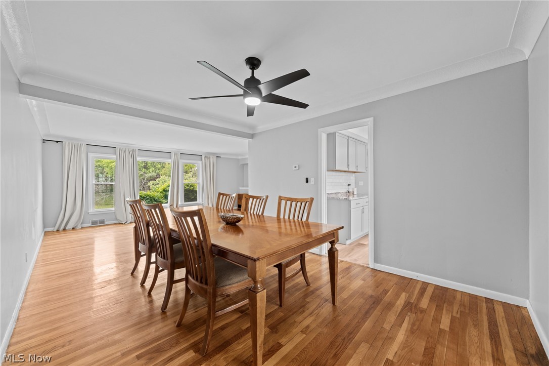 Dining room with a wooden table, hardwood floors, and curtained windows overlooking greenery.