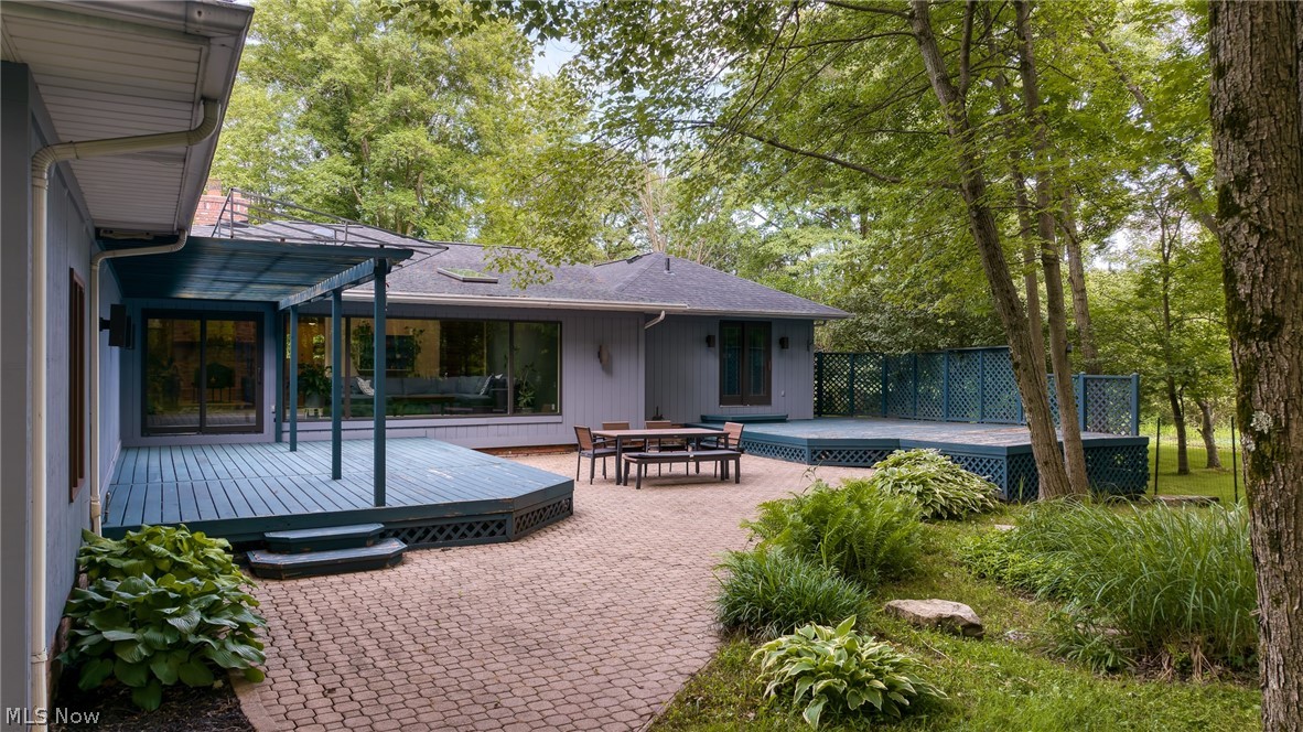 Home’s rear patio with covered blue deck, brick pavers, outdoor dining set, and mature trees.