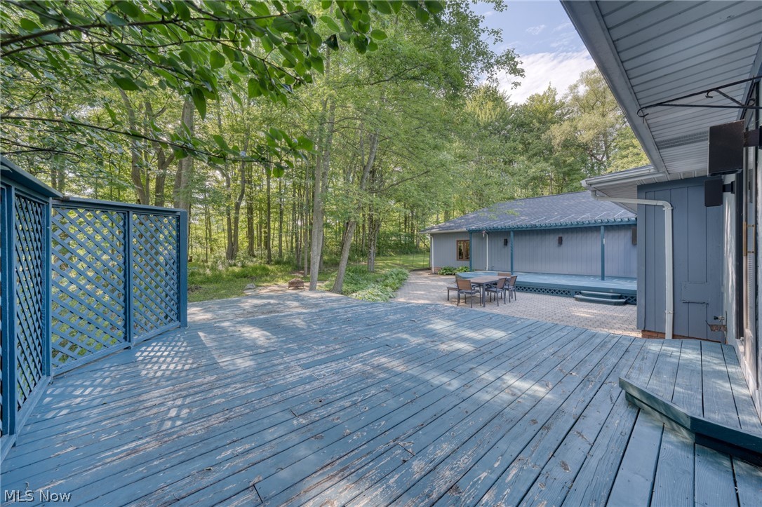 Blue-gray backyard deck beside a swimming pool and outdoor dining table, bordered by leafy trees.