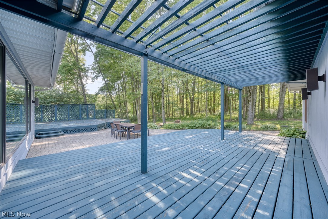 Blue deck beneath an open-slat pergola, with outdoor dining furniture and woods beyond.