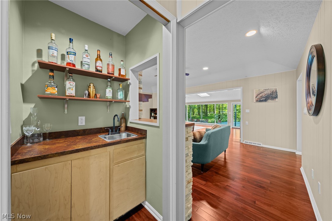 Wet bar with open bottle shelves and a sink beside a living room with hardwood floors.