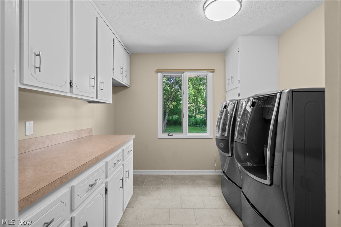 Laundry room with white cabinetry, a long countertop, dark front-loading washer and dryer, and a garden-view window.