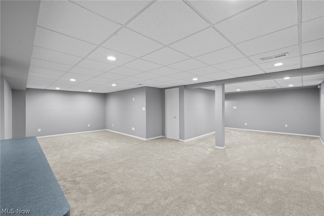 Carpeted basement with gray walls, a suspended ceiling, support columns, and a closed door.