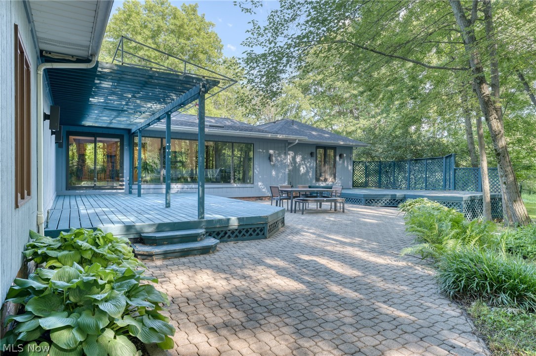 Modern home’s raised deck and pergola overlook a paved patio with outdoor dining furniture and mature trees.