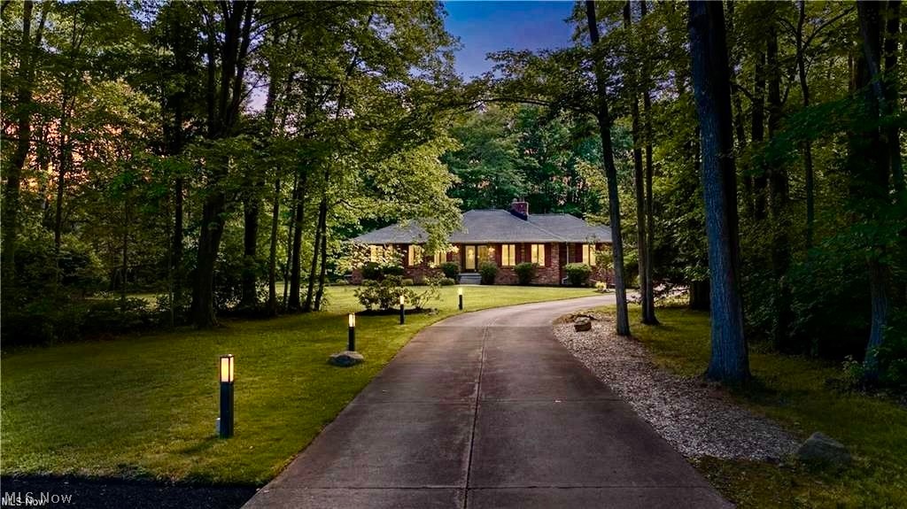 Brick home with glowing windows at the end of a curved driveway beneath mature trees.