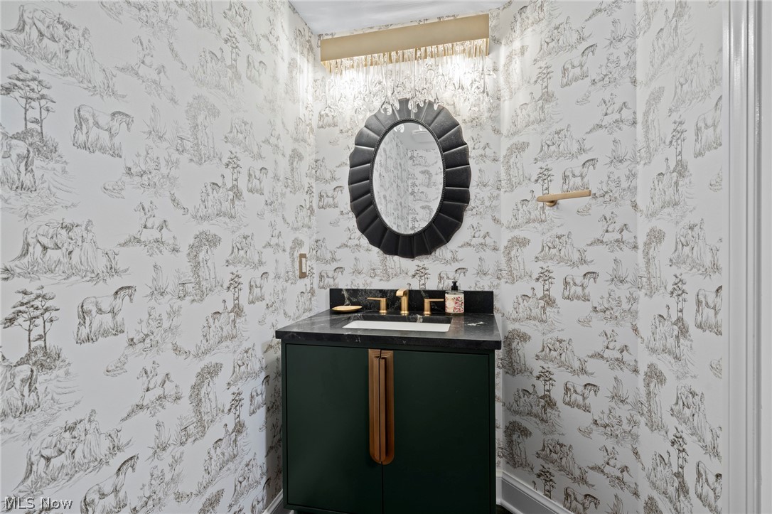 Powder room with toile wallpaper, a dark green vanity, brass fixtures, and a black oval mirror.
