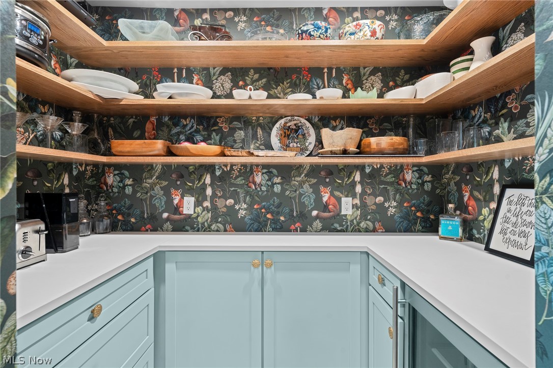 Pantry with pale blue cabinetry, open wood shelves, displayed dishes, and fox-and-bird floral wallpaper.