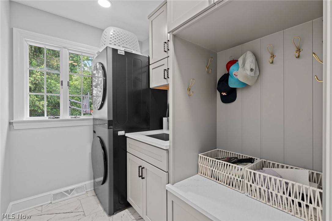 Laundry mudroom with stacked black washer and dryer, white cabinetry, brass hooks, and a window overlooking greenery.