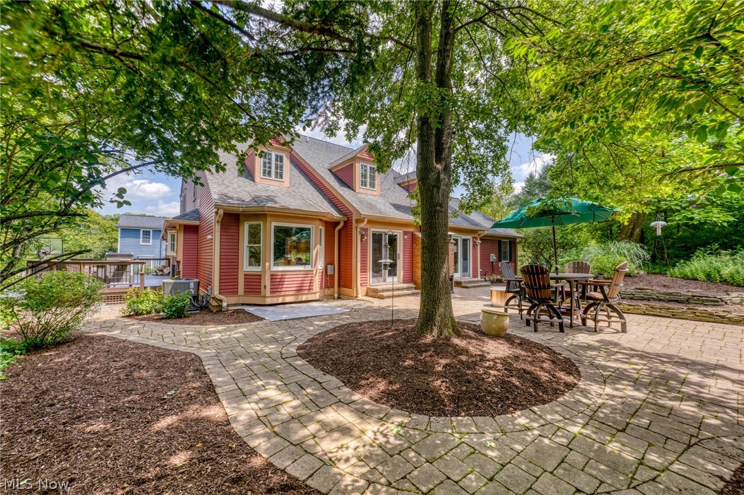 Red-sided house with a stone patio, outdoor dining set, green umbrella, and large shade tree.