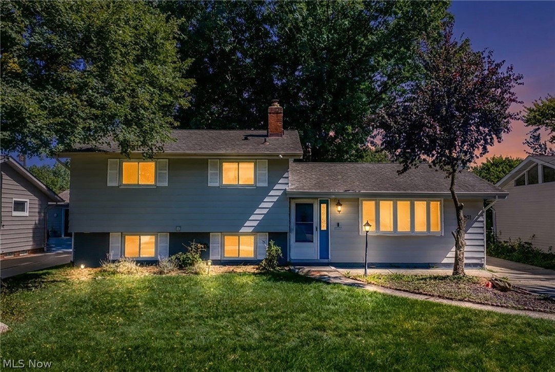 Blue-gray split-level house at dusk with glowing windows, mature trees, and a front lawn.