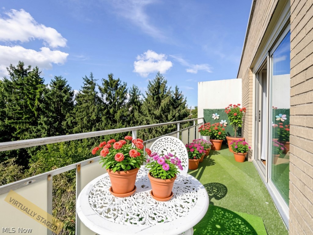 Balcony with a white patio table, potted flowers, and evergreen trees beyond the railing.