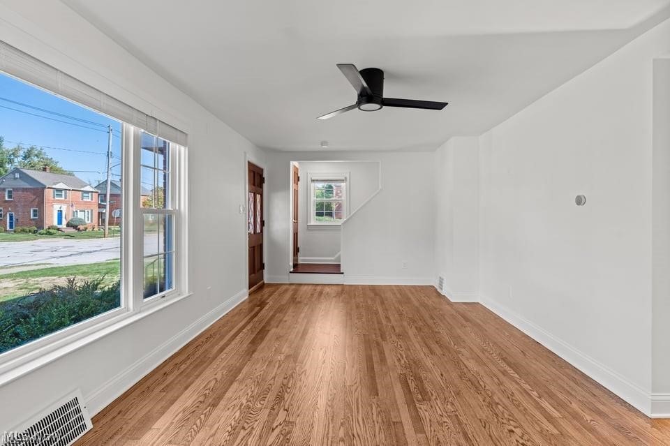 Empty living room with hardwood floors, large front windows, and a black ceiling fan.
