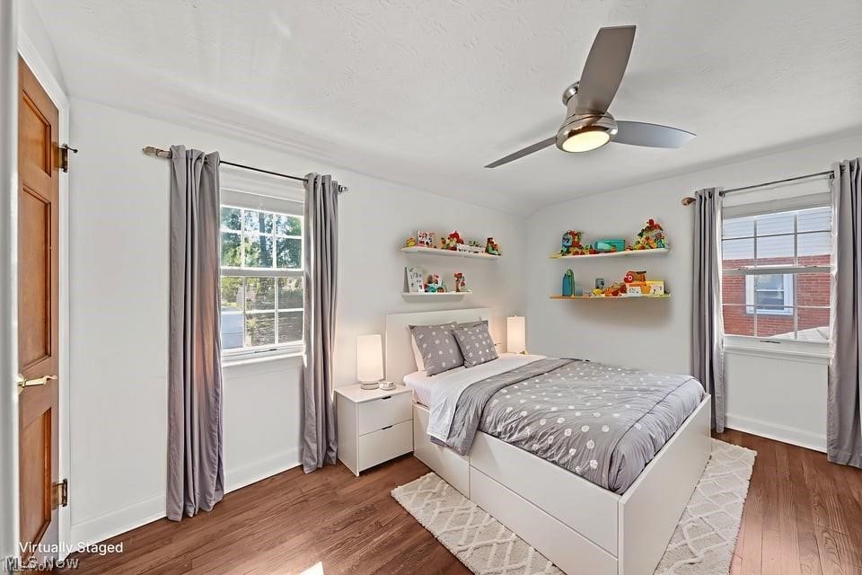 Virtually staged bedroom with a gray bed, toy-filled shelves, two windows, and wood flooring.
