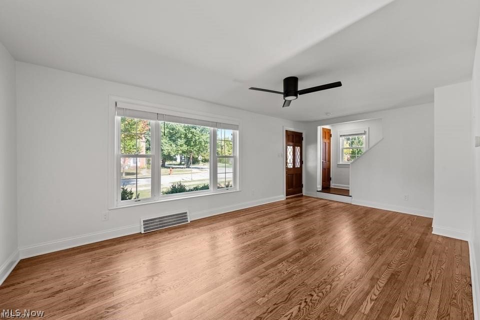 Empty living room with polished hardwood floors, a wide front window, and a black ceiling fan.