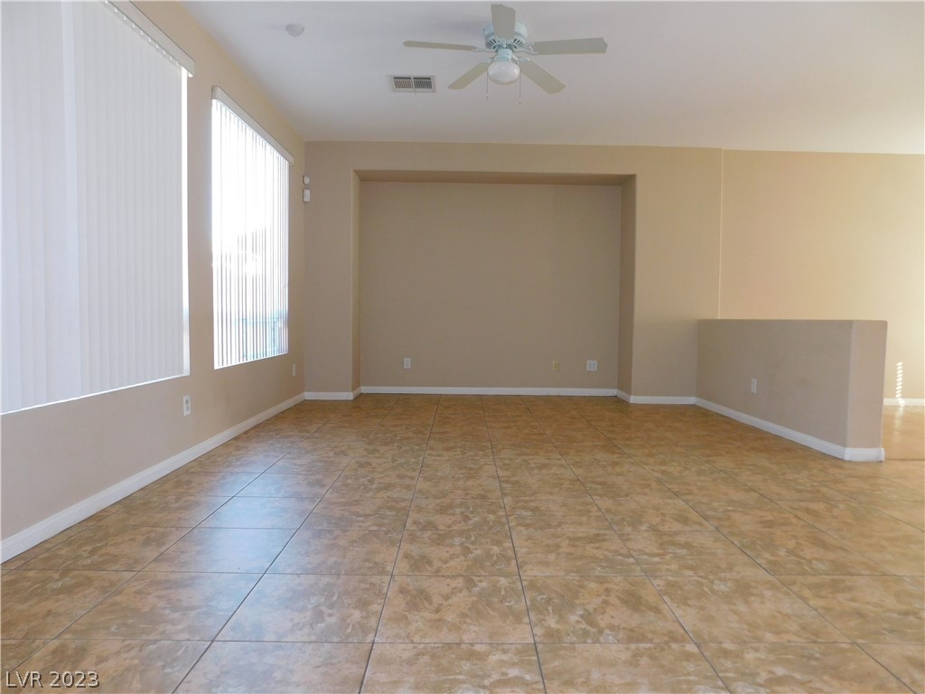 Empty living area with tan tile floors, large window blinds, and a ceiling fan.