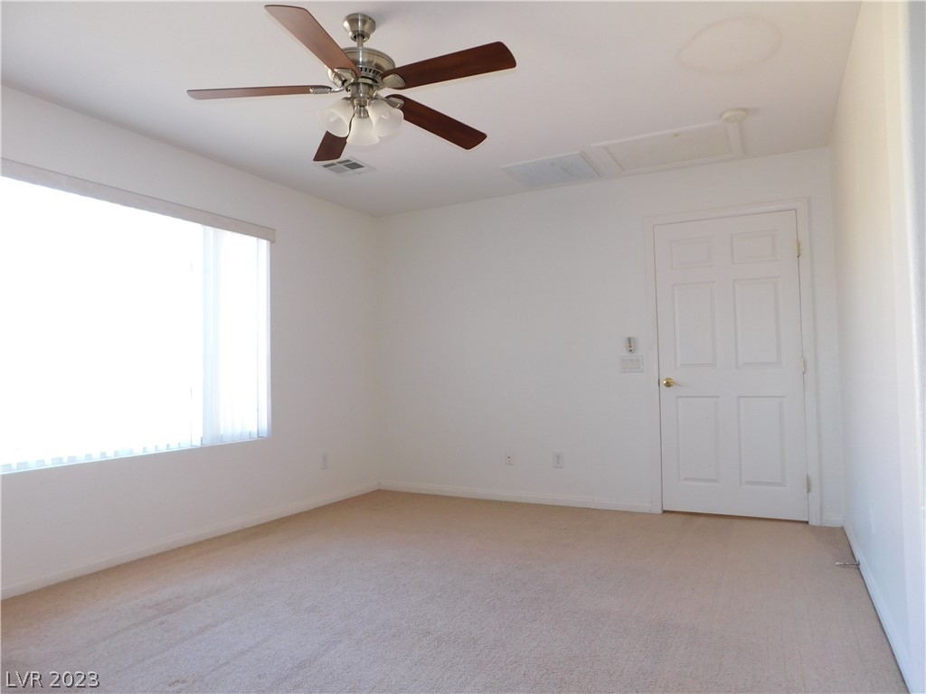 Empty bedroom with beige carpet, a large window with vertical blinds, and a dark-bladed ceiling fan.