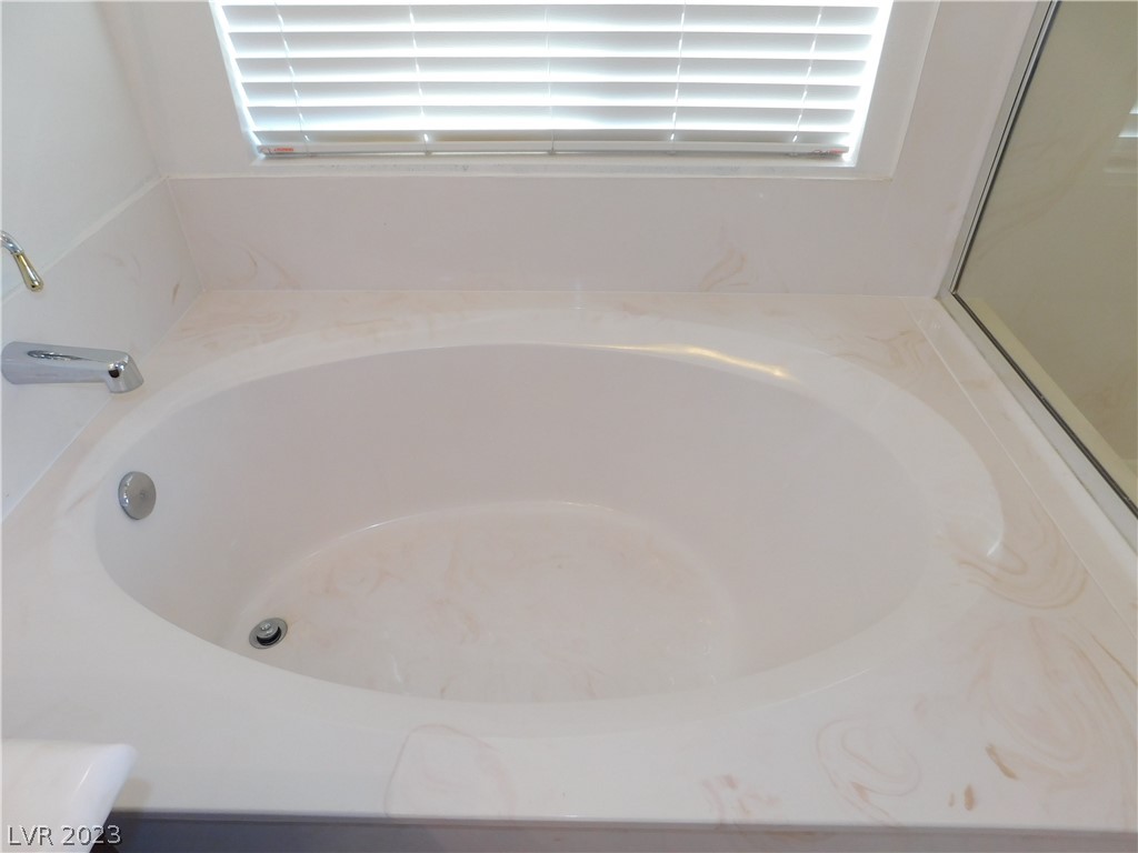 Oval bathtub beneath a shuttered window, with a pale marble-patterned surround and glass shower enclosure.