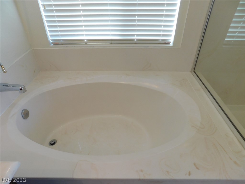 Oval soaking tub with a cream marble-patterned surround beneath plantation shutters.