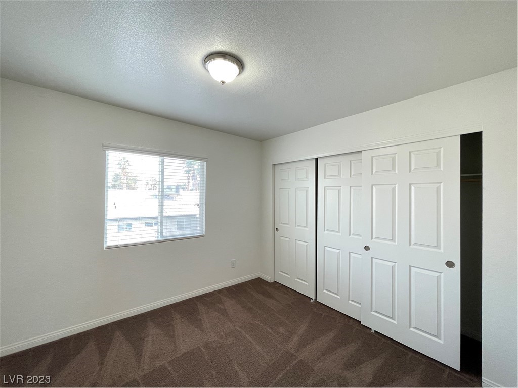 Empty bedroom with brown carpet, a window with blinds, and open closet doors.