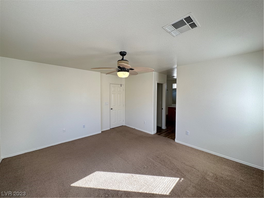 Empty carpeted bedroom with a ceiling fan, paneled door, and sunlight across the floor.